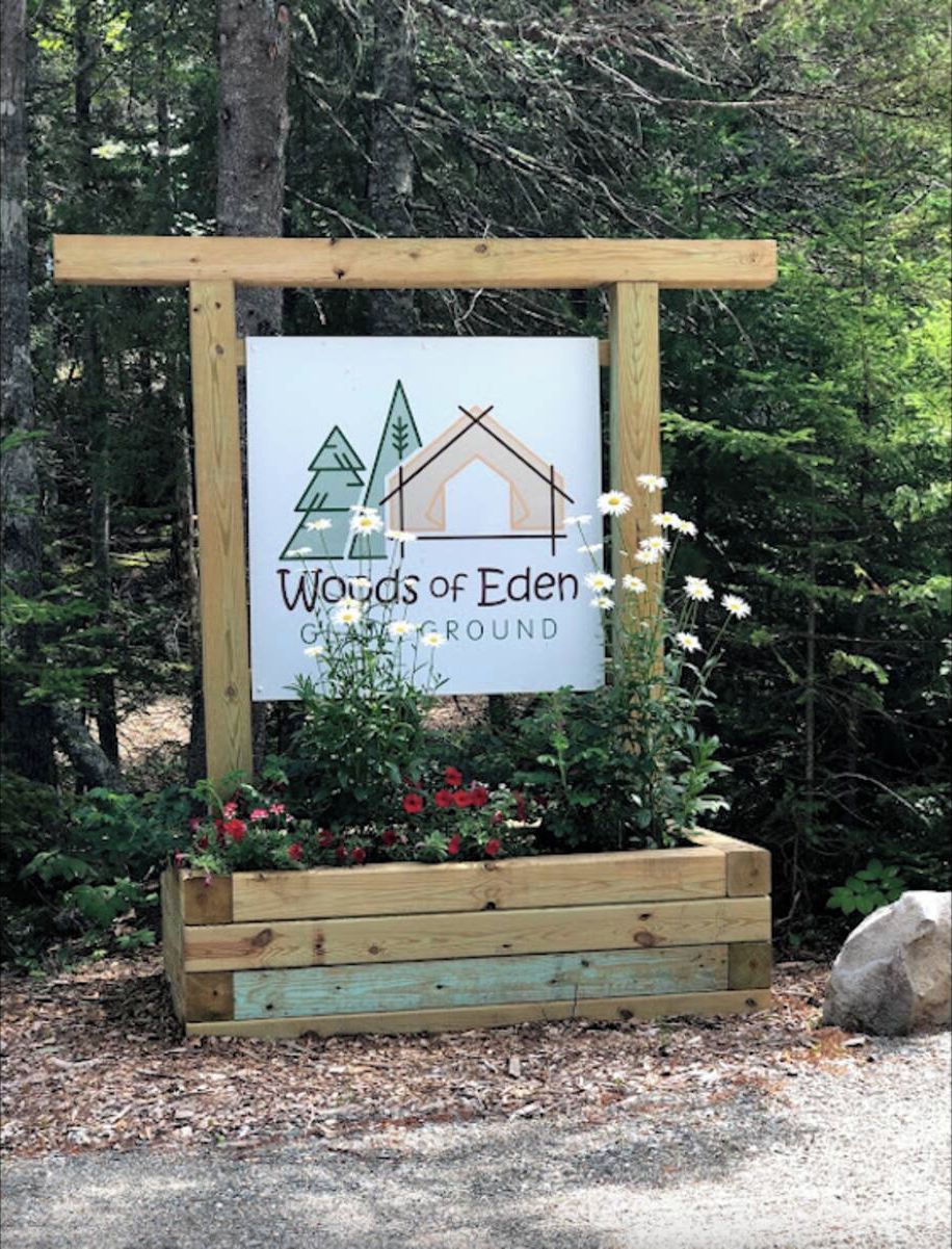 A sign for 'Woods of Eden Campground' with a wooden structure and a planter with flowers.
