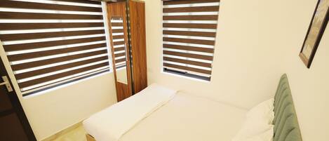 A bedroom with a bed, two windows with blinds, a door, and a wall-mounted picture frame.