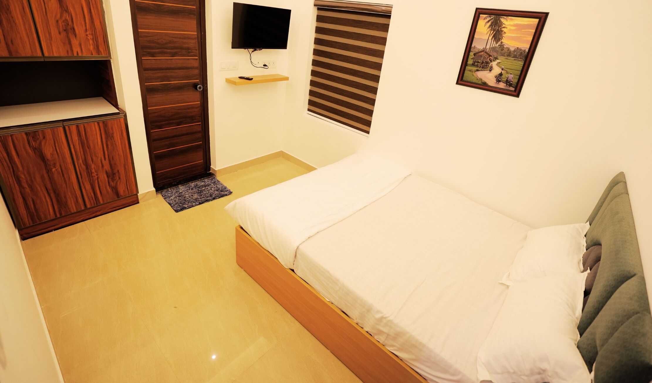 A bedroom with a bed, a wooden wardrobe, a television, and a painting on the wall.