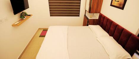 A neatly made bed with white linens, a red headboard, a mounted TV, a small plant, and a window with blinds.