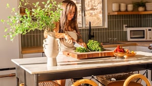 Una cucina con un piano di lavoro in marmo, una pianta in vaso, un tagliere con verdure fresche e una ciotola di frutta.