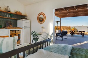 A cozy room with a bed, a small table, and chairs. There's a kitchen area with a refrigerator and shelves. Outside, there's a patio with a table and chairs.