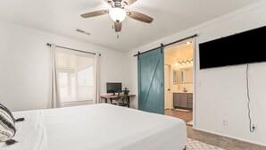 A bedroom with a bed, a desk, a ceiling fan, and a bathroom visible through a sliding door. - The Farm House | Pool&Spa | 5 min to JT Park (Twentynine Palms)