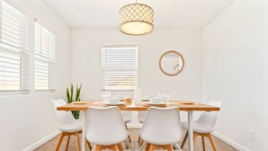 A dining area with a wooden table, white chairs, a hanging light fixture, and a window with blinds. - The Farm House | Pool&Spa | 5 min to JT Park (Twentynine Palms)