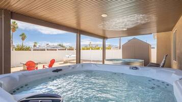 A hot tub with a view of a white fence and palm trees, under a wooden roof structure.