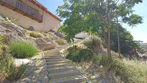Uma escadaria de pedra serpenteia por uma colina, rodeada por vegetação e um edifício com varanda.