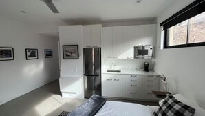 Een moderne slaapkamer met een bed, nachtkastje, plafondventilator, raam met zonwering en een kitchenette met koelkast, magnetron en spoelbak.