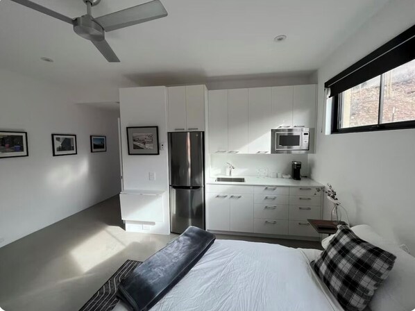 A modern bedroom with a bed, a bedside table, a nightstand, a ceiling fan, a window with blinds, and a kitchenette with a refrigerator, microwave, and sink.