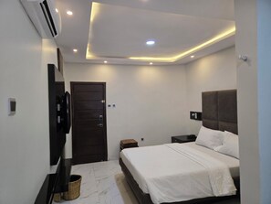A modern hotel room with a large bed, a flat-screen TV mounted on the wall, a dark wooden door, and a ceiling with recessed lighting. - NEKST Hotel (Ibadan)