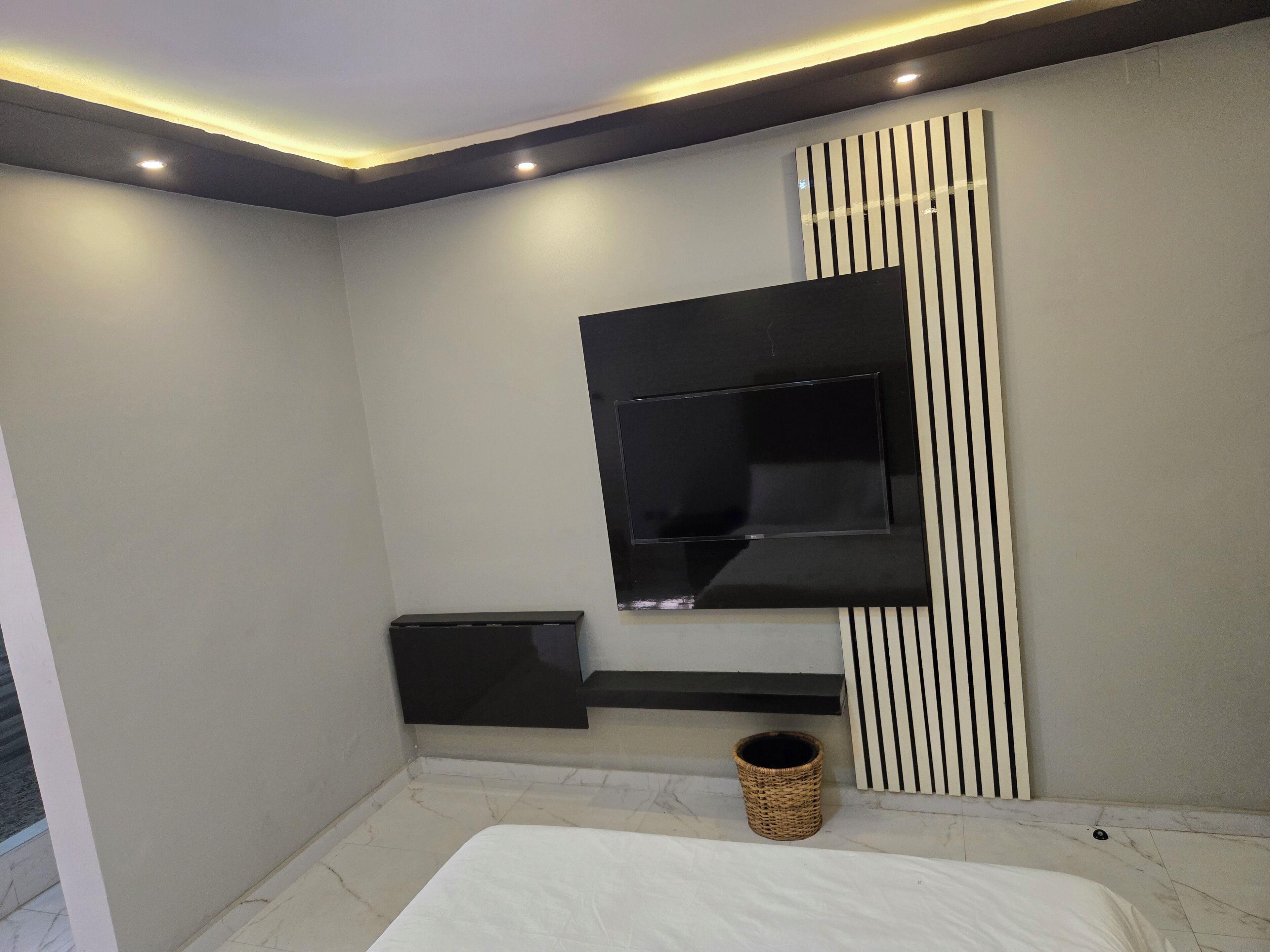 A modern bedroom with a large bed, a mounted TV, a small storage unit, and a decorative wall panel.