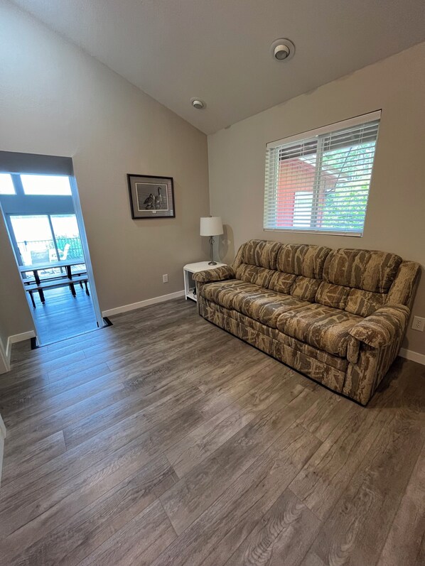 A spacious living room with a patterned sofa, a small side table with a lamp, a framed picture on the wall, and a window with blinds.