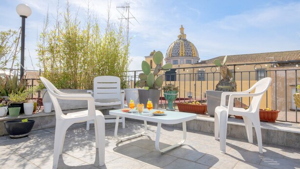 An outdoor patio with white plastic chairs and a table, surrounded by potted plants and a building with a dome in the background. - mimi terrace (Naples)