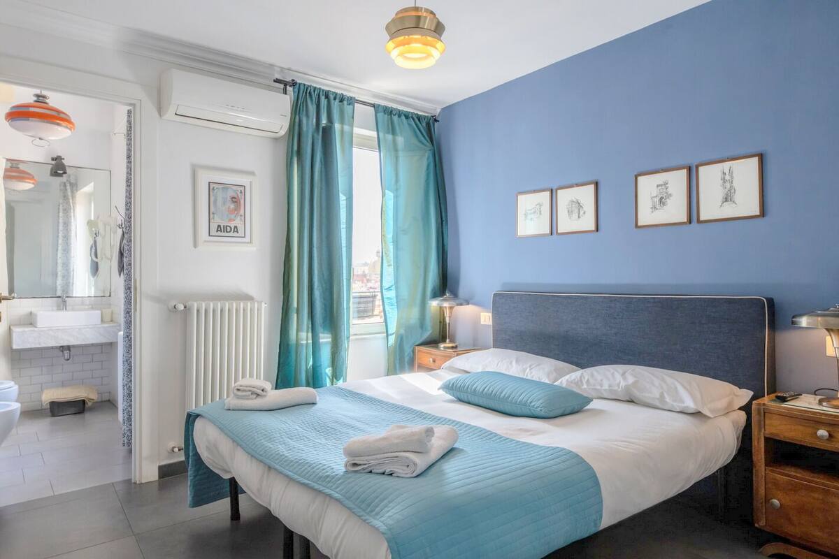 A bedroom with a bed, a nightstand, a bedside lamp, a window with curtains, a wall with framed pictures, and a bathroom visible through an open door.