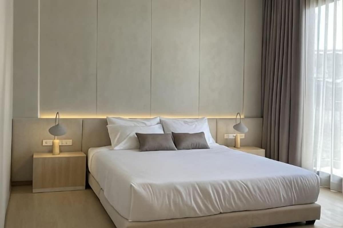 A modern bedroom with a large bed, two bedside tables with lamps, and a view of the outdoors through a window.
