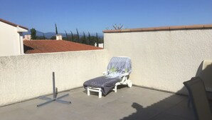 A rooftop terrace with a single lounge chair, a metal table, and a view of distant buildings and trees. - Apartment in a haven of peace 10 mm from the Mediterranean Sea. (Sorède)