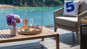 A balcony with a wicker chair, a table with a bottle, glasses, and a basket of snacks.