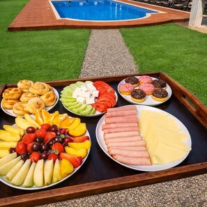 A tray with various food items including fruits, cheeses, and pastries, placed outdoors with a pool in the background. - Blue Escape (Nafplio)