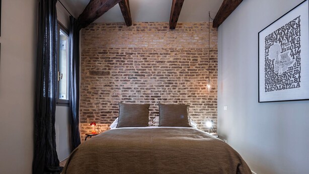 A bedroom with a brick wall, a bed with a brown cover, a framed picture on the wall, and a window with curtains. - Cá delle Capriate, an intimate retreat at Cà d'Oro (Venezia)