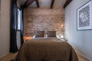 Un dormitorio con pared de ladrillo, una cama con cubierta marrón, un cuadro enmarcado en la pared y una ventana con cortinas.