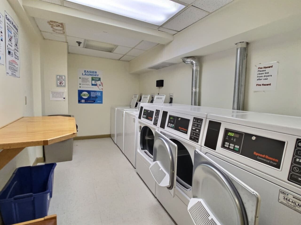 A laundry facility with multiple commercial dryers, a wooden bench, and a blue waste bin.