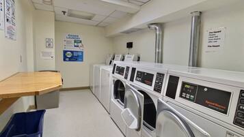 A laundry facility with multiple commercial dryers, a wooden bench, and a blue waste bin.