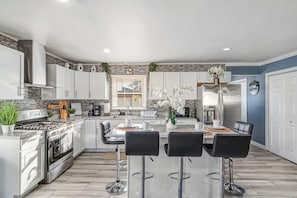 A modern kitchen with white cabinets, stainless steel appliances, a central island with bar stools, and decorative plants.