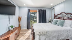 Un dormitorio con cama, cortinas, televisor, planta y vistas al exterior.
