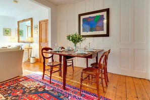 A dining area with a wooden table set for four, surrounded by chairs, a patterned rug, and a framed abstract painting on the wall.
