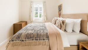 Ein Schlafzimmer mit Bett, Nachttisch, einem Fenster mit Vorhängen, einer Pendelleuchte und einem Wandbild.