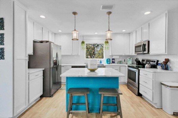 Cocina moderna con armarios blancos, electrodomésticos de acero inoxidable, una isla central con taburetes y luminarias colgantes.