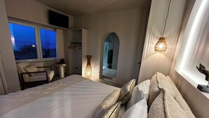 Quarto de hotel com uma cama grande, uma mesinha de cabeceira, uma cadeira, uma janela e um banheiro visível por um arco.