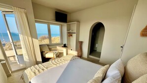 Quarto com cama grande, uma cadeira, mesinha e vista para o mar através de uma janela.