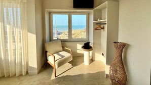 Quarto com uma janela grande, uma cadeira, uma mesa pequena, uma televisão e um vaso decorativo.
