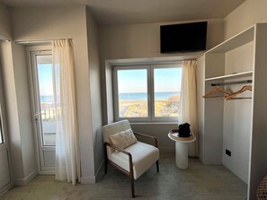 A room with a window view of the beach, a chair, a small table with a coffee pot, and a TV mounted on the wall.