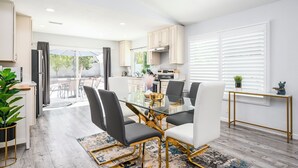 Uma cozinha moderna com armários brancos, uma sala de jantar com mesa de vidro e cadeiras pretas, e vista para o exterior por grandes janelas.