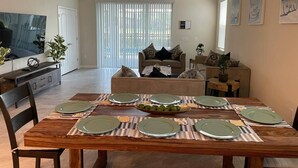 A dining area with a wooden table set for six, a sofa in the background, and a television on the left. - Your Home by the Orlando Parks (Davenport)