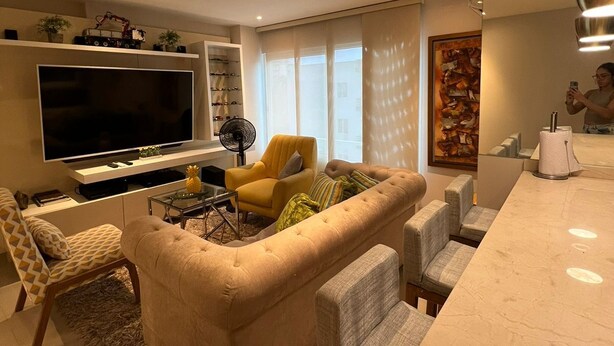Una sala de estar moderna con un televisor de pantalla plana grande, un sillón amarillo, un sofá capitonado y una zona de comedor con una mesa de mármol.