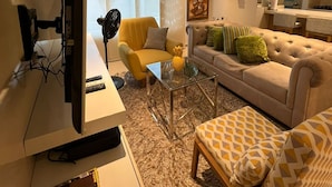 Una sala de estar moderna con un televisor de pantalla plana, un sillón amarillo, un sofá color beige y un reposapiés estampado.