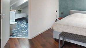 Une chambre avec un plafond en pente, un lit recouvert d’une couvre-lit à motifs, un banc et une vue sur une pièce adjacente dont le sol est décoré de motifs bleus et blancs.