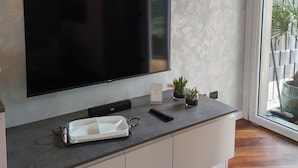 Un salon moderne avec un téléviseur à écran plat fixé au mur, une console blanche sur laquelle repose une télécommande, une petite plante en pot et un vase décoratif.