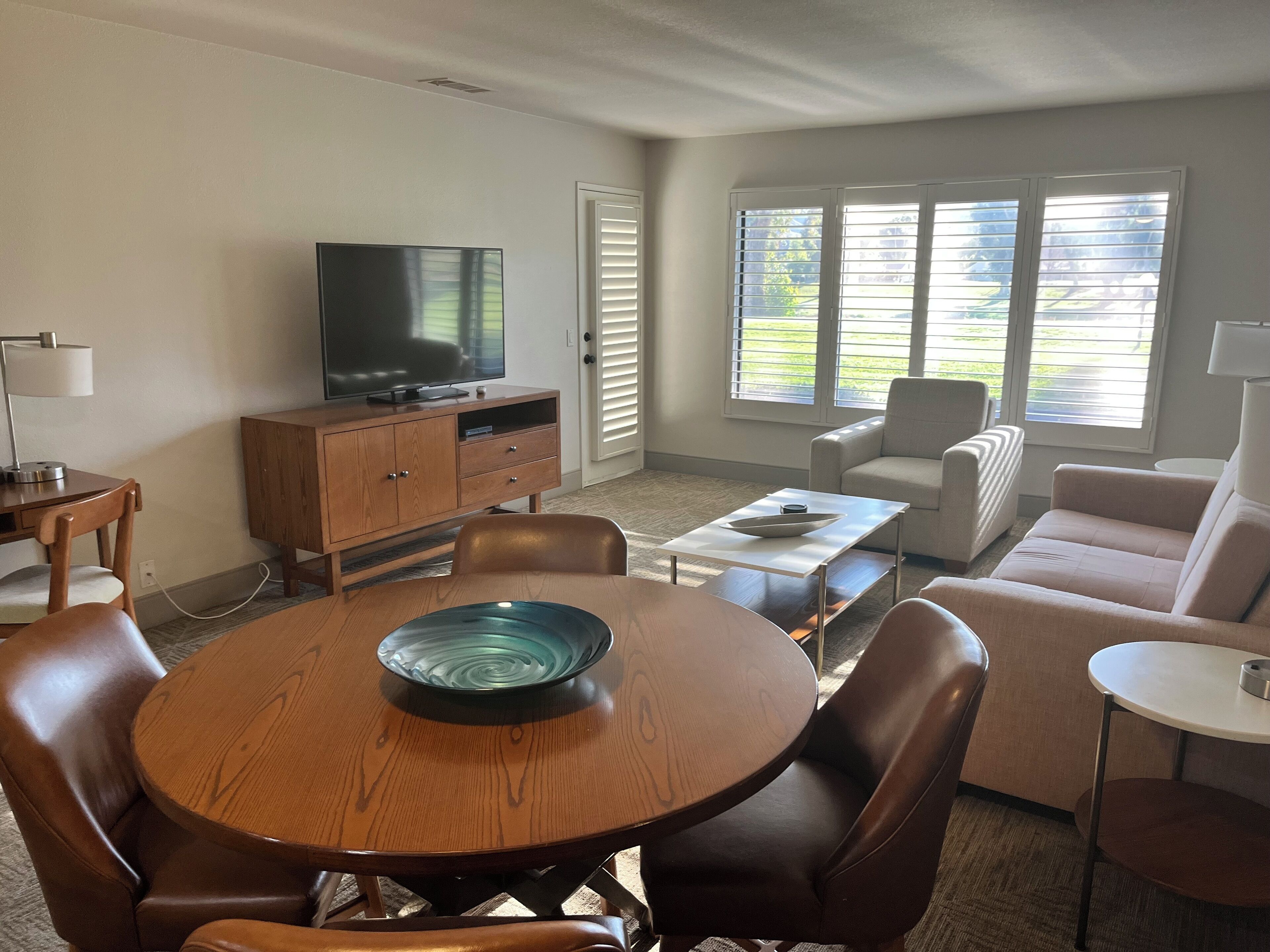 Een woonkamer met een houten eettafel, lederen fauteuils, een bank, een televisie op een houten kast en een raam met zonwering.