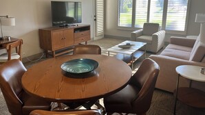Ein Wohnzimmer mit Esstisch aus Holz, Ledersesseln, einer Couch, einem Fernseher auf einer Holzkommode und einem Fenster mit Jalousien.