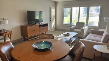 Een woonkamer met een houten eettafel, lederen fauteuils, een bank, een televisie op een houten kast en een raam met zonwering.