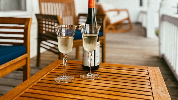 Una mesa de madera con dos copas de vino blanco y una botella de vino en un porche.