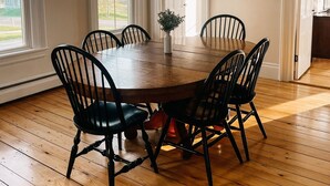 Comedor con mesa de madera rodeada por seis sillas, una lámpara de araña y vistas al exterior a través de ventanas amplias.