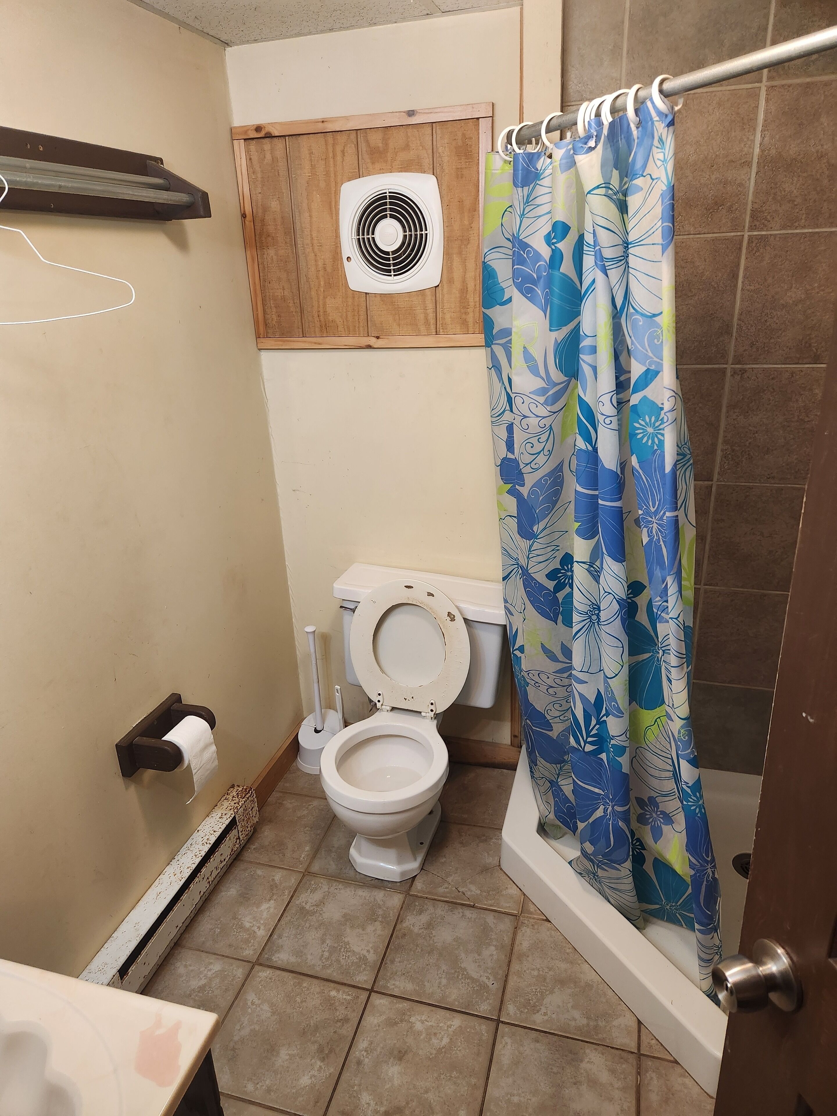 A bathroom with a toilet, a shower curtain, a fan, and a towel rack.