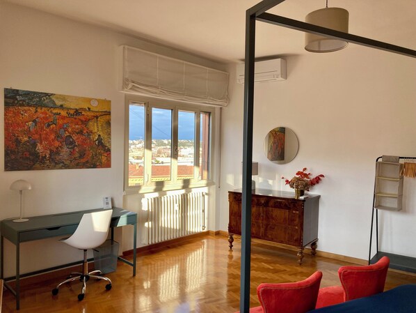 A modern living room with a large window, a desk, a chair, a sofa, and a radiator.