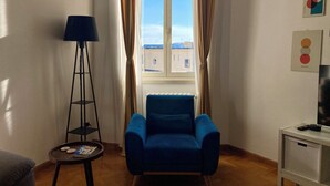 Une pièce avec un fauteuil bleu, une lampe sur pied, une petite table avec un magazine et une fenêtre avec des rideaux.