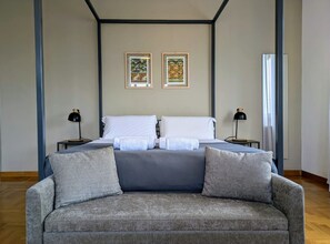 A bedroom with a bed, two nightstands, a grey sofa, and two framed artworks on the wall.