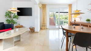 A modern living room with a marble coffee table, a sofa, a TV mounted on the wall, and a dining area with a wooden table and chairs. - Casa Torremuelle II – Townhouse with private Pool & BBQ near Playa Bonita (Benalmádena)